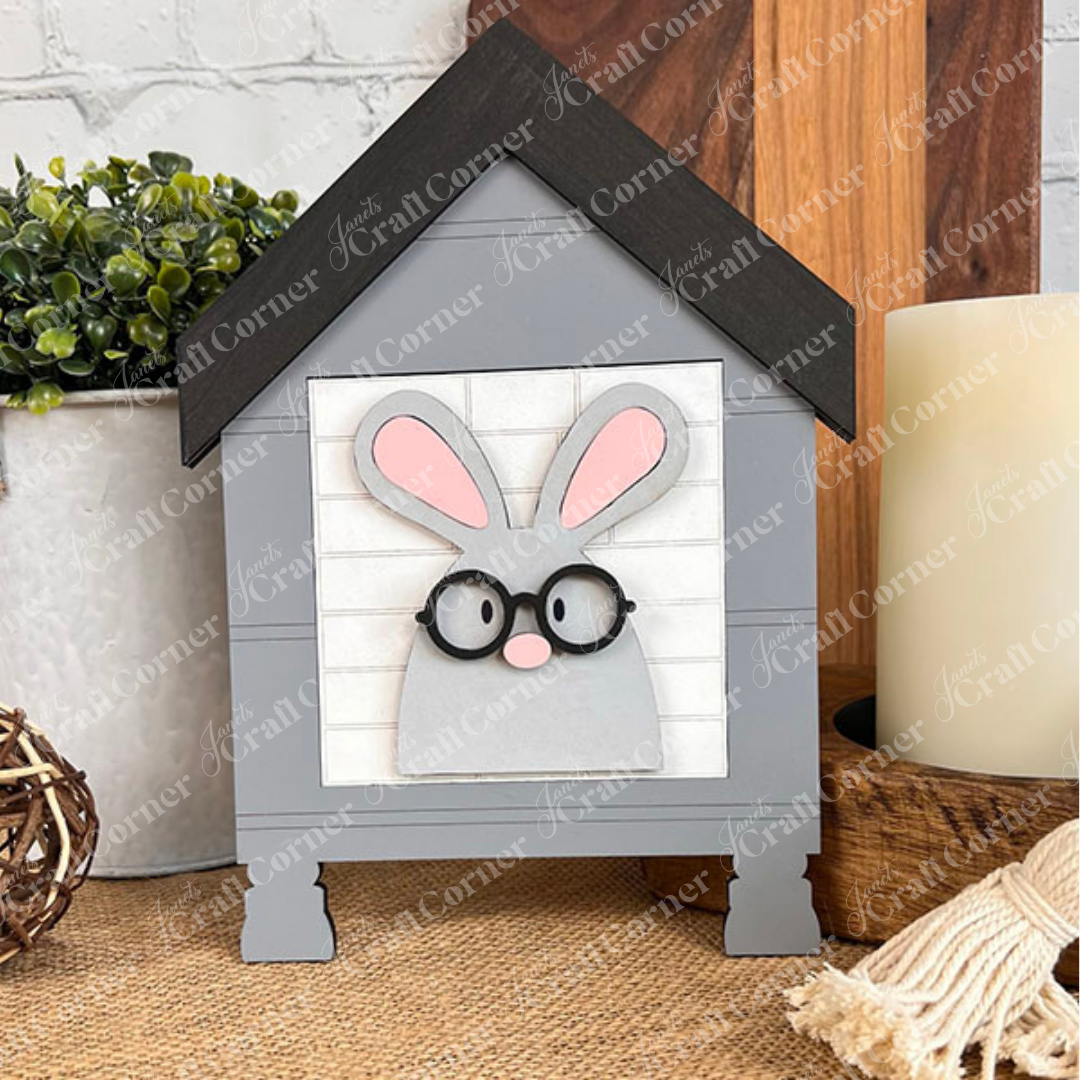 The Easter Bunny Squares by Janet's Craft Corner is a small, house-shaped gray wood sign with a bunny face, glasses, and pink ears—ideal for tiered tray decor alongside plants or woven balls.