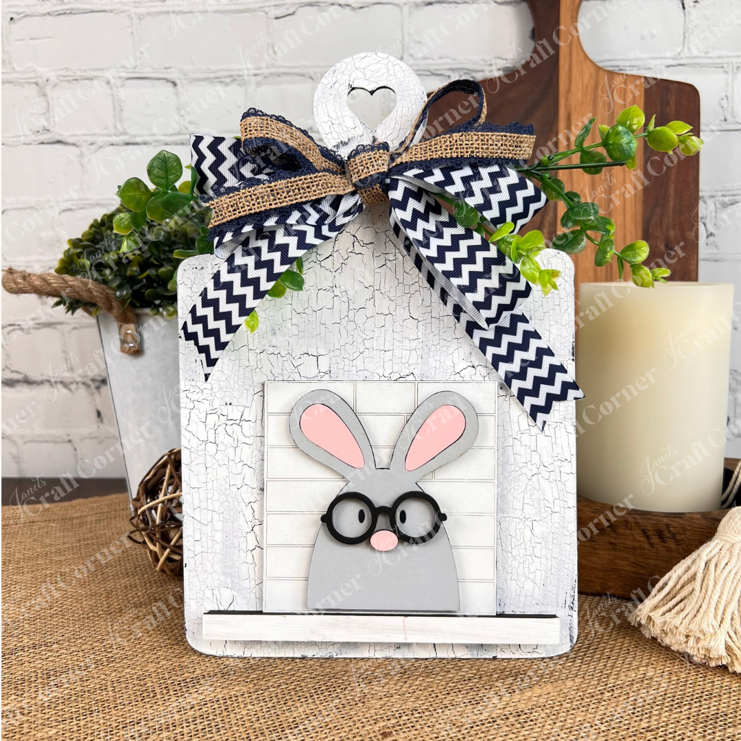 Janet's Craft Corner Easter Bunny Squares: A cute gray bunny with glasses and pink ears sits before faux greenery and a white brick backdrop, accented by a chevron ribbon—perfect for tiered tray decor or Swappable Square Displays.