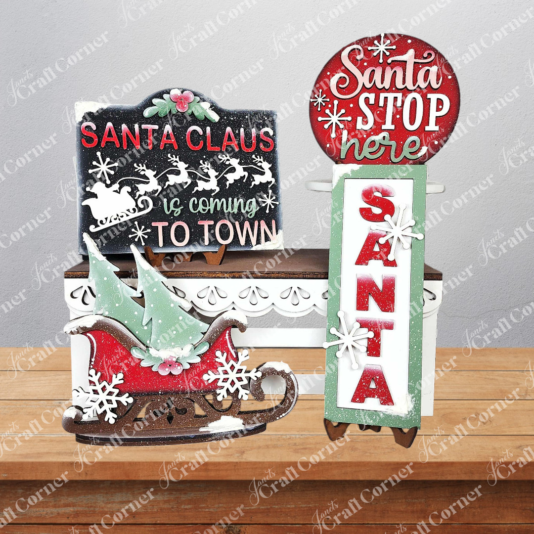 The North Pole Decor Set by Janet's Craft Corner features four festive Christmas decorations: “Santa Claus is coming to town” and “Santa Stop Here” signs, a red sleigh with gifts, and a vertical “Santa” sign with snowflakes.