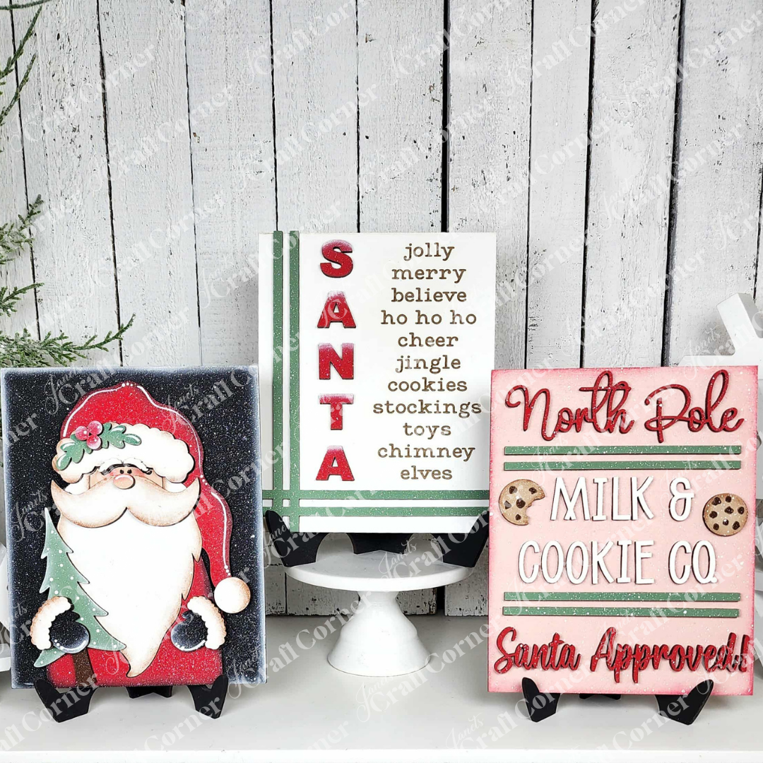 Janet's Craft Corner North Pole Switchable Farmhouse Signs include a Santa figurine sign, a vertical "SANTA" sign with Christmas words, and a pink "North Pole Milk & Cookie Co." sign—festive DIY holiday home decor or craft kit addition.