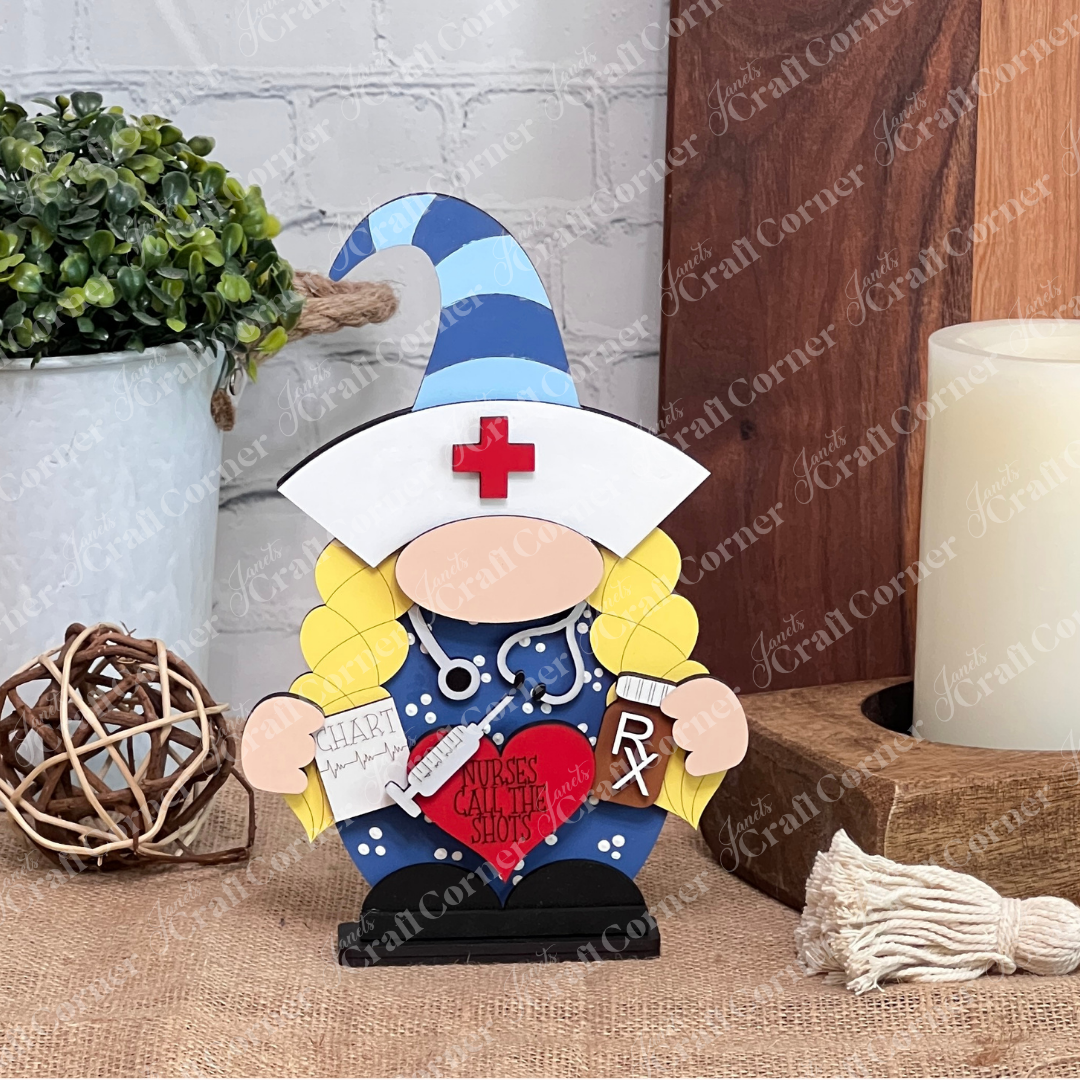 Introducing the Nurse Gnome from Janet's Craft Corner, a delightful wooden figurine featuring a blue hat and a stethoscope. This DIY kit also comes with a chart and syringe accessory. The gnome holds a red heart inscribed with "Nurses Call the Shots," making it an ideal addition to farmhouse decor. Perfectly suited for display on tabletops surrounded by decorative plants and wooden accents.