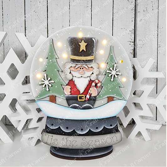 Bring festive cheer to your home with the Nutcracker Light-Up Snow Globe Shelf Sitter from Janet’s Craft Corner. This DIY home decor craft kit features a nutcracker Santa holding a candy cane, flanked by two green trees and surrounded by white snowflakes. The base displays the phrase “’Tis the Season,” while small lights illuminate Santa, making this charming set of 1 a delightful holiday shelf accent.