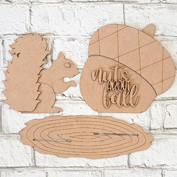 The "Nuts About Fall Shelf Sitter" from Janet's Craft Corner is a DIY home decor kit that includes unique wooden craft pieces such as a squirrel silhouette, a large acorn with the engraved message "nuts about fall," and an oval slice reminiscent of a tree ring. When displayed against a white brick background, these elements add charming character to any space.