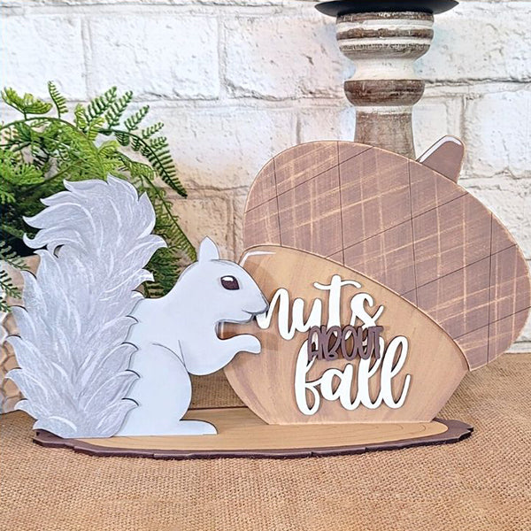 The "Nuts About Fall Shelf Sitter" from Janet's Craft Corner is a delightful DIY home decor kit that features a hand-painted decorative sign with a white squirrel next to a large acorn. The sign, complete with the phrase "Nuts About Fall," is beautifully set against a potted fern and white brick wall backdrop, making it an ideal addition for craft enthusiasts looking to enhance their home decor charm.