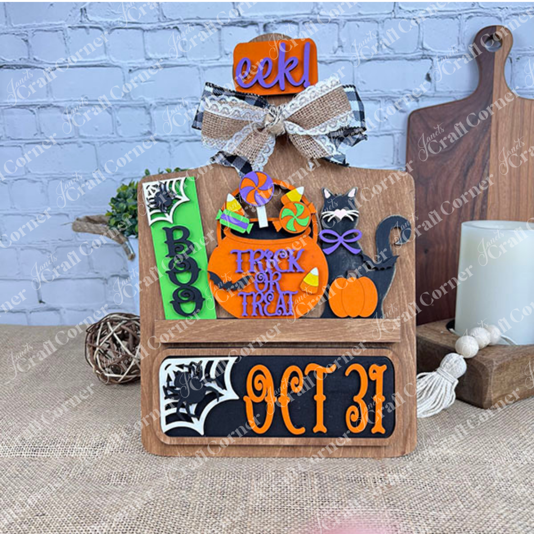 Janet's Craft Corner offers a DIY interchangeable home decor kit featuring a rustic wooden base with inserts displaying "BOO," "EEK!" signs, a black cat, candy, pumpkins, "Trick or Treat" message, "OCT 31" in bold letters, and decorative bows.