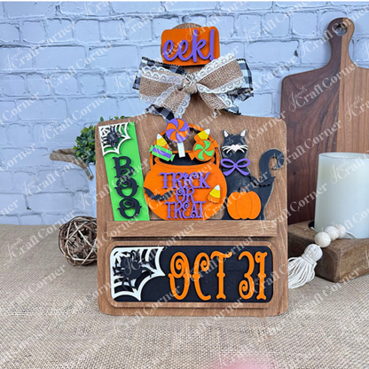 Janet's Craft Corner offers a DIY interchangeable home decor kit featuring a rustic wooden base with inserts displaying "BOO," "EEK!" signs, a black cat, candy, pumpkins, "Trick or Treat" message, "OCT 31" in bold letters, and decorative bows.