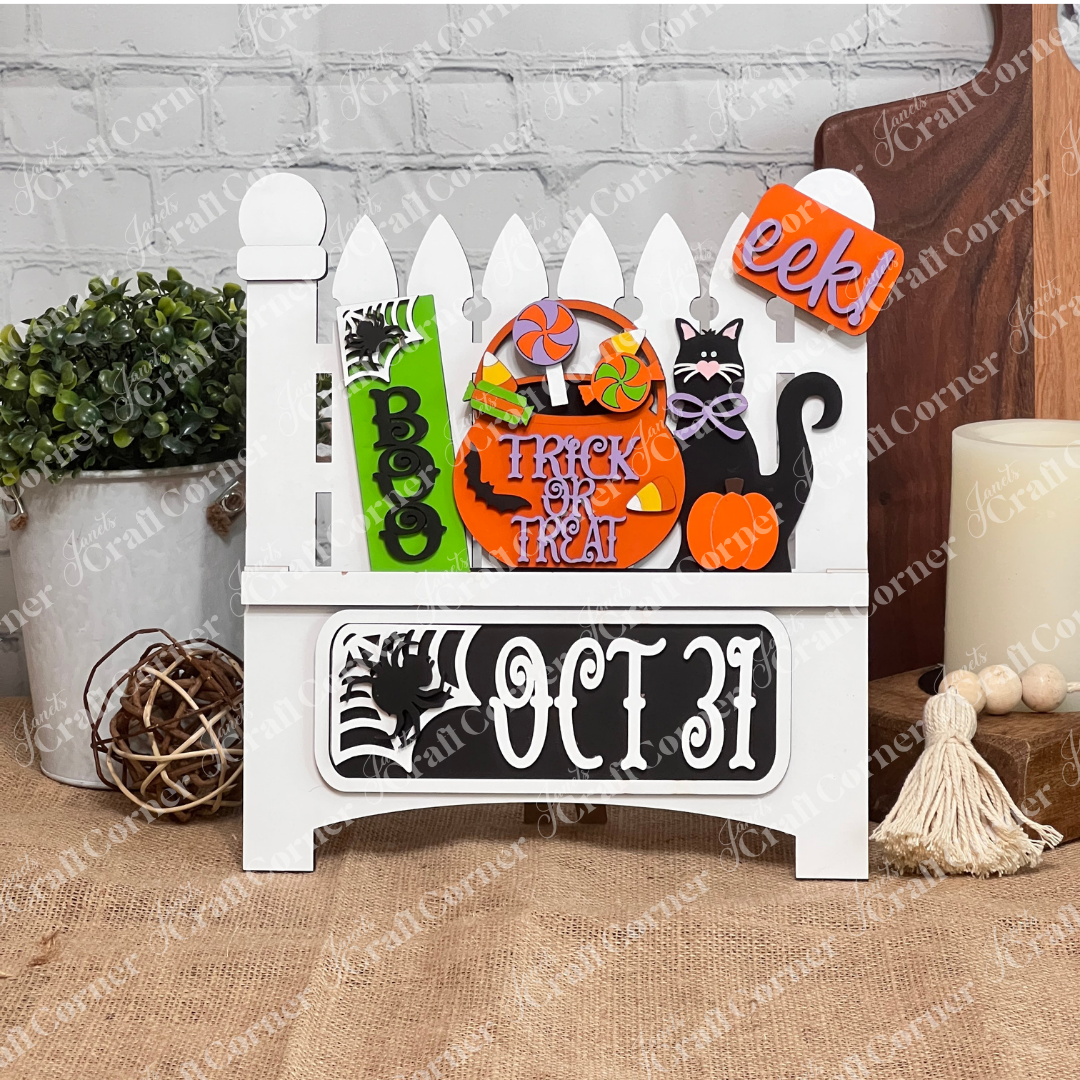 Janet's Craft Corner offers a Garden Fence with Insert, featuring a wooden Halloween-themed design adorned with "Boo," "Trick or Treat" signs, a black cat, pumpkin, lollipops, and spider web against a white brick wall—perfect for festive home decor.