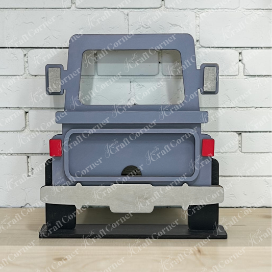 Introducing the Off Road Truck Interchangeable Base DIY home decor craft kit from Janet’s Craft Corner—a wooden decor piece designed to resemble the rear view of a Jeep, painted gray with vibrant red tail lights. Displayed in front of a crisp white brick wall on a rustic wooden surface, this versatile accent is an ideal addition to any farmhouse-style seasonal display.
