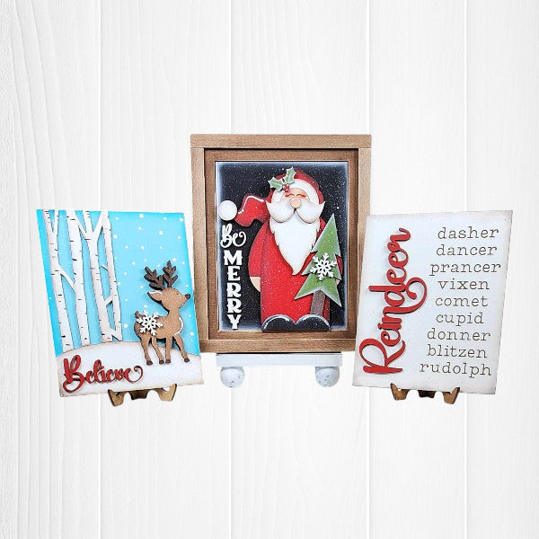 Janet's Craft Corner offers the Farmhouse Frame for Interchangeable Signs, a DIY home decor kit featuring a festive farmhouse style. It includes wooden plaques with designs: reindeer by birch trees with "Believe," Santa with "Be Merry," reindeer names with "Reindeer," and a small pine tree.