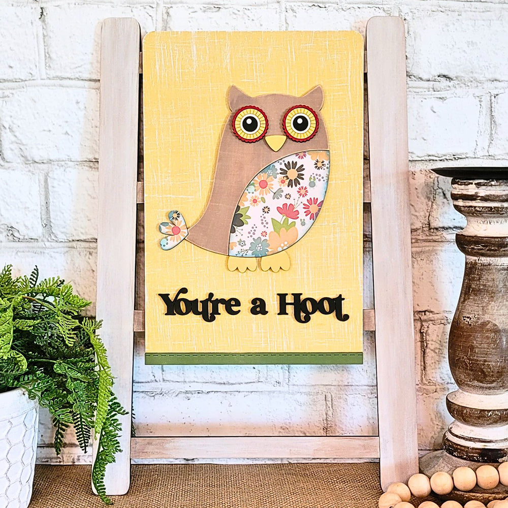 A delightful addition to your home, the "You're a Hoot" Tea Towel from Janet's Craft Corner showcases an owl with floral-patterned wings set against a sunny yellow backdrop. The towel features the playful phrase "You're a Hoot" and can be displayed on its rustic wooden stand, providing a whimsical accent to any space.