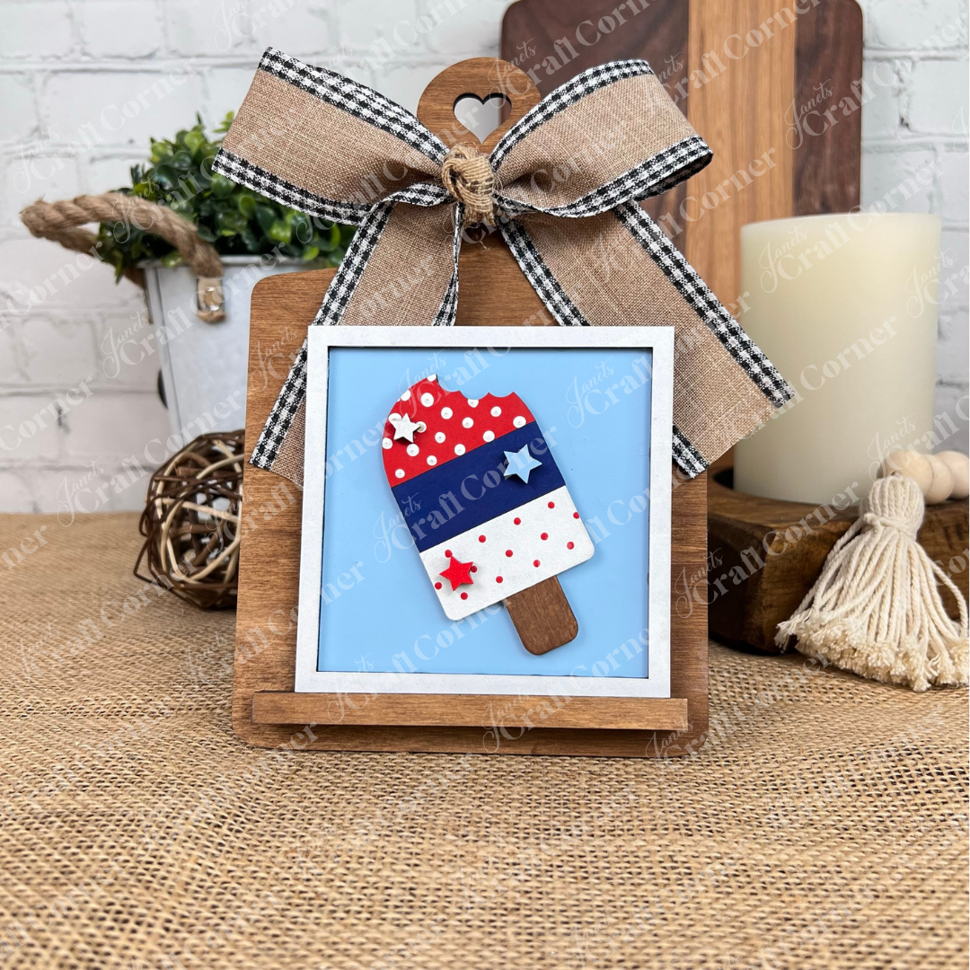 Display Janet's Craft Corner’s "Patriotic Squares" DIY seasonal home decor craft kit—a framed red, white, and blue popsicle with stars—on a wooden easel with a burlap and plaid bow for charming rustic Independence Day décor.