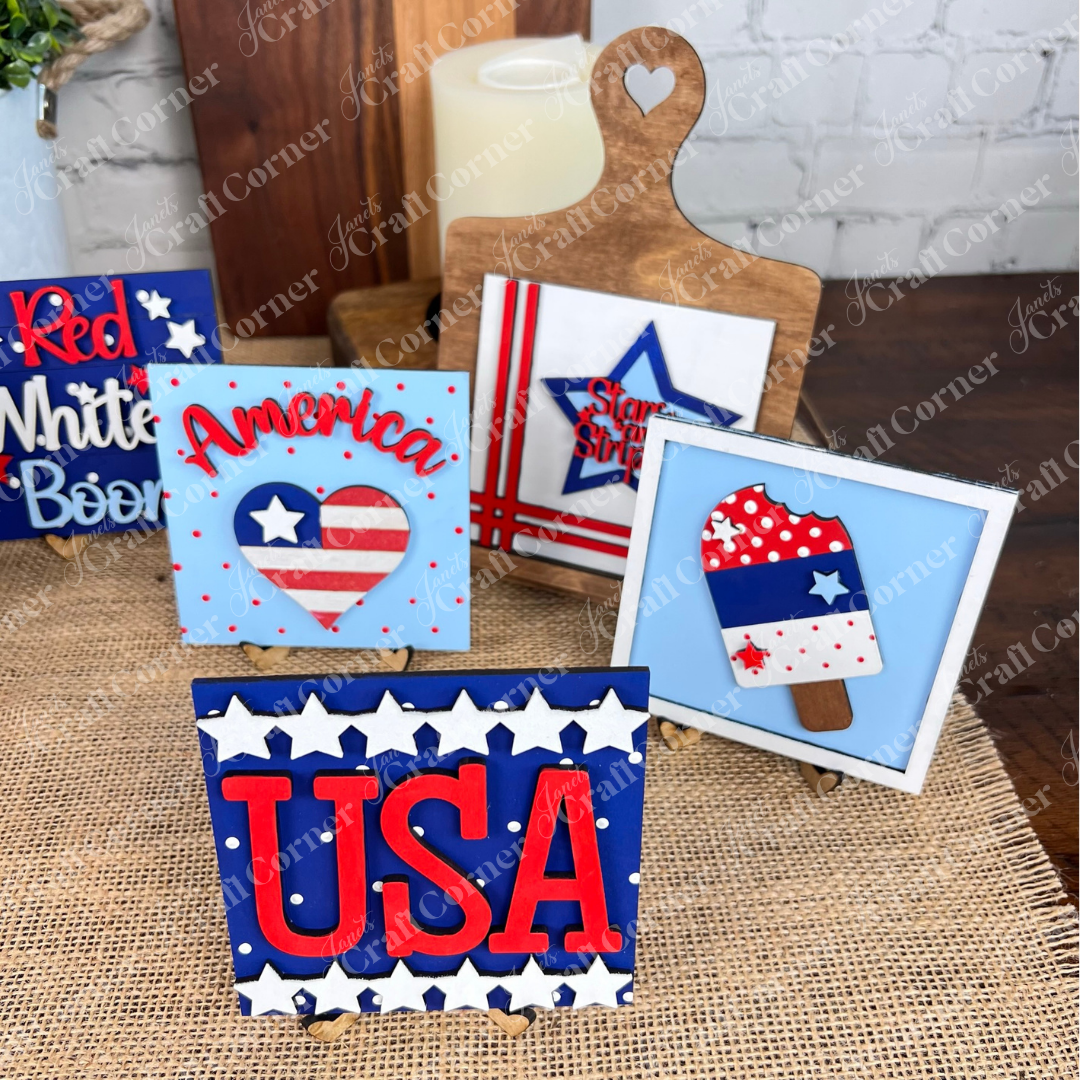 Janet’s Craft Corner’s Patriotic Squares DIY craft kit features wooden signs for Independence Day, including “USA,” “America” with a heart, a stars-and-stripes popsicle, and a heart-topped cutting board in festive red, white, and blue.