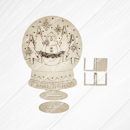 Laser-cut wooden snow globe craft kit featuring a cheerful snowman, trees, and “IT’S FREEZIN SEASON” text, shown with flat oval and square stand pieces on a white background. Perfect as DIY holiday decor for your home or to pair with Janet’s Craft Corner Penguin Snowglobe Shelf Sitter – DIY home decor craft kit (set of 1).