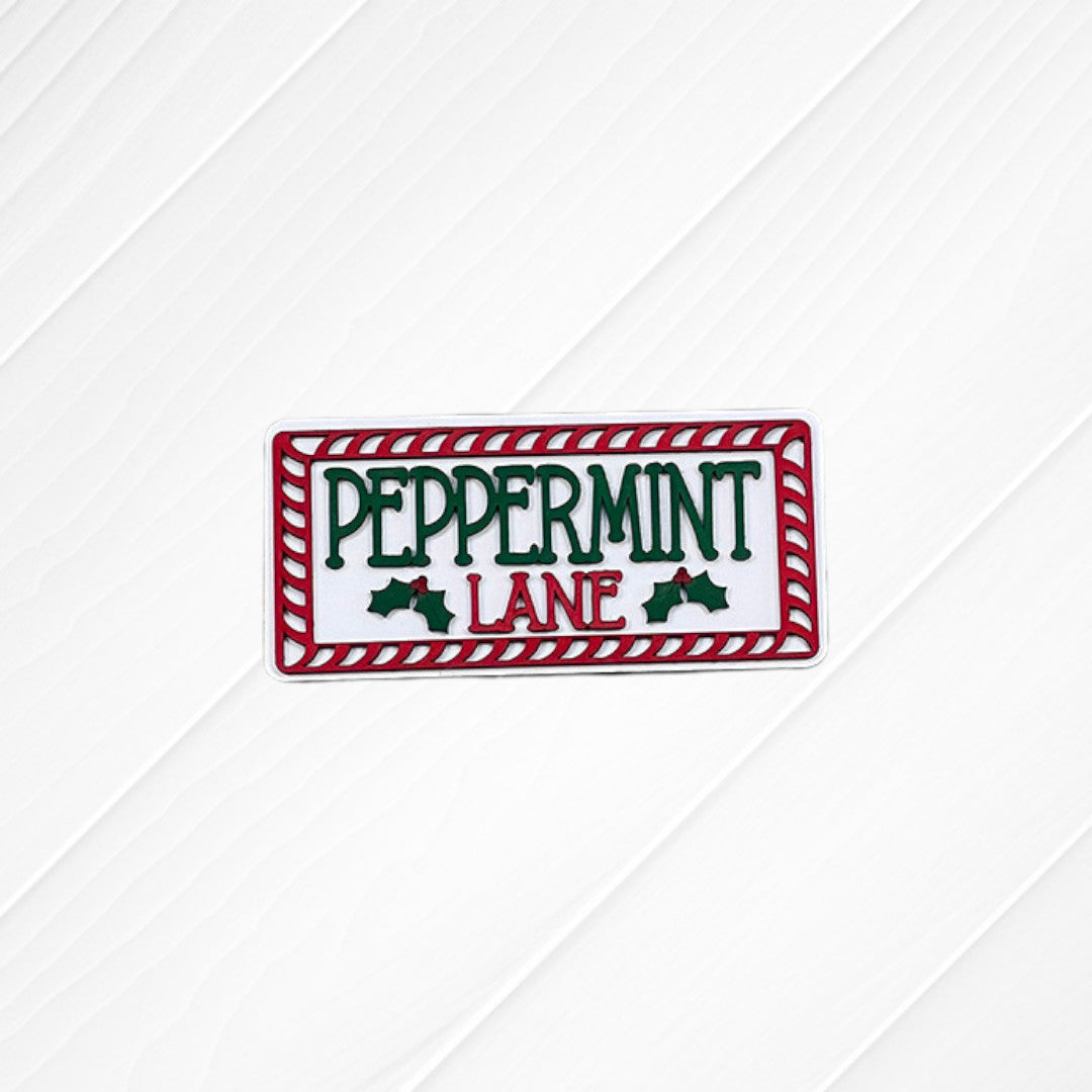 A festive sign from Heartland’s Peppermint Mocha Mini Shelf Sitters collection features “PEPPERMINT LANE” in green and red letters, bordered by a red and white candy cane pattern and accented with holly leaves. Painted on a white wooden background, this charming piece is perfect for holiday home decor or as an accent to your tiered tray display.
