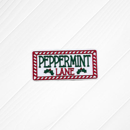 A festive sign from Heartland’s Peppermint Mocha Mini Shelf Sitters collection features “PEPPERMINT LANE” in green and red letters, bordered by a red and white candy cane pattern and accented with holly leaves. Painted on a white wooden background, this charming piece is perfect for holiday home decor or as an accent to your tiered tray display.
