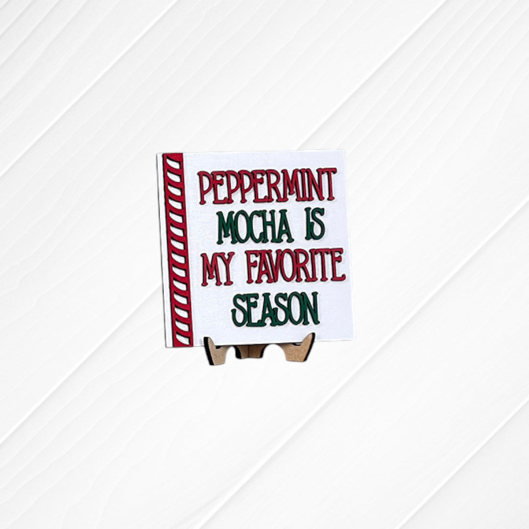 A Heartland Peppermint Mocha Mini Shelf Sitter—painted in festive red and green with the phrase “Peppermint Mocha Is My Favorite Season”—is displayed on a wooden stand against a white textured background, making it the perfect piece to add holiday cheer to your tiered tray kit or seasonal decor display.