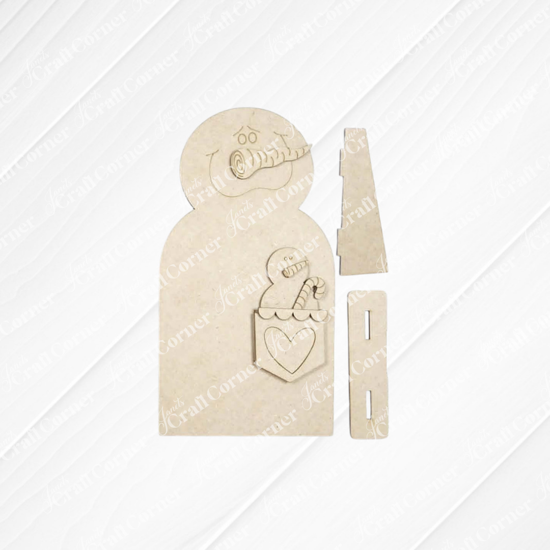 Bring a touch of winter charm to your home with the Pocket Snowman Shelf Sitter DIY craft kit by Janet’s Craft Corner. This unassembled kit includes a wooden snowman featuring a carved face, scarf, and a pocket filled with candy canes and a heart, along with two rectangular pieces—all set against a light background. Perfect for creating your own festive shelf decor!.