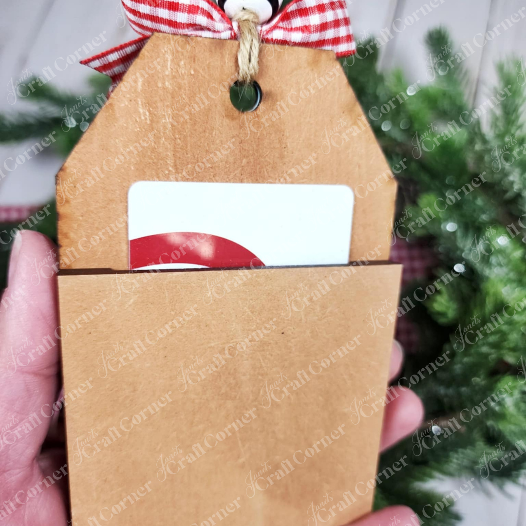 A hand holds a brown kraft paper envelope with a red and white striped ribbon, revealing a Forest Friends Ornaments – DIY craft kits (set of 5) gift card from Janet's Craft Corner. Blurred greenery is in the background.