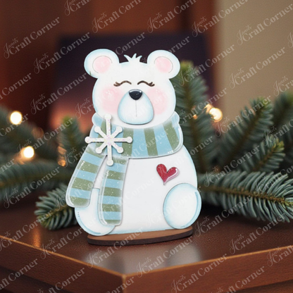 A Polar Bear Shelf Sitter from Janet's Craft Corner—featuring a white bear with pink cheeks, closed eyes, a green striped scarf, and a red heart—is displayed on wood by pine branches and string lights. DIY decor kit; choice of 2.