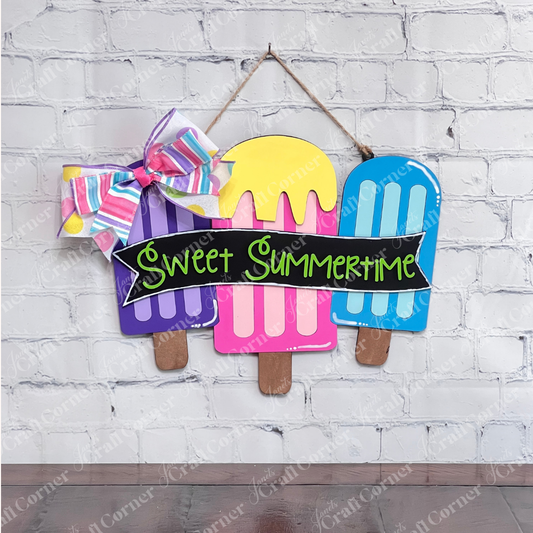 The Sweet Summertime Door Hanger by Janet's Craft Corner showcases three vibrant ice cream popsicles in shades of purple, pink, and orange, accentuated with lively lettering. It features a charming red and orange polka dot bow on the top left corner against a textured wooden background, making it perfect for home décor or a delightful DIY kit project.