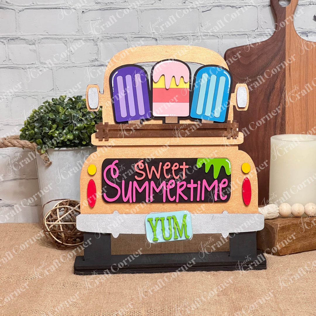 Add a charming touch to your home décor with Janet's Craft Corner's DIY home decor kit, featuring the Popsicle Interchangeable Insert. This decorative piece showcases a copper truck with vibrant popsicles and "Sweet Summertime" elegantly written in pink and orange above a teal "YUM" license plate. It’s perfect for placing next to a potted plant on a wooden surface.
