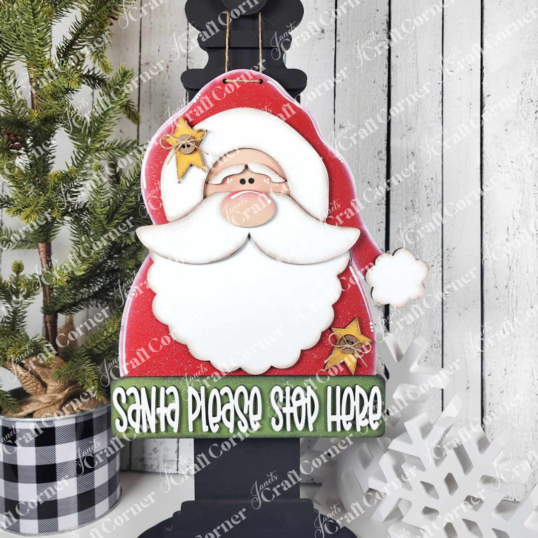 Janet's Craft Corner Primative Santa Door Hanger in a red suit and white beard hangs on a black post with a "Santa please stop here" sign—ideal holiday decor, pictured with a small pine tree and snowflake background.