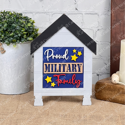 Janet's Craft Corner’s Military Squares is a small house-shaped sign for tiered tray decor. It reads "Proud MILITARY Family" in blue, beige, and red with yellow stars—perfect for displaying on a table or shelf.