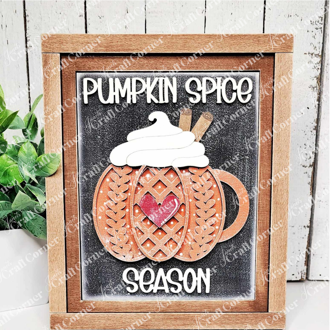 Immerse yourself in the cozy atmosphere with Janet's Craft Corner's Pumpkin Spice Season Interchangeable Sign. This DIY home decor craft kit features a decorative framed sign with a wooden mug, highlighted by a lattice pumpkin design, whipped cream, cinnamon sticks, and is adorned with a red heart. Embrace the spirit of "Pumpkin Spice Season" as text accompanies the design while a plant adds charm on the left.