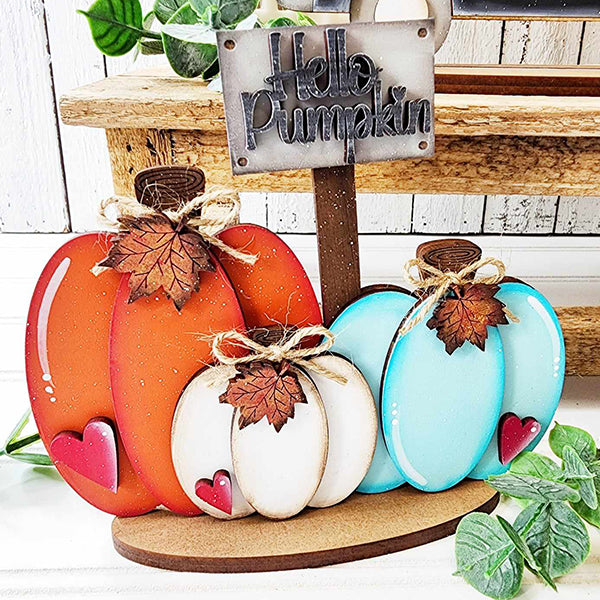 The Pumpkins Mini Shelf Sitter by Janet's Craft Corner is a DIY home decor kit that includes decorative wooden pumpkins in orange, white, and blue. Adorned with leaf accents and twine bows, these hand-painted pieces are designed to sit on a wooden shelf next to a "Hello Pumpkin" sign. Some pumpkins also feature small red heart accents for an extra touch of love.