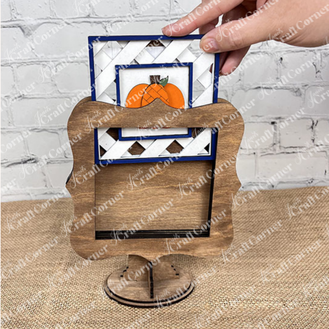 A hand places a pumpkin-themed card into the Heartland Drop In Swappable Squares Holder DIY Craft Kit (Set of 1), perfect for home décor. The scene shows a wooden surface and a white brick wall in the background.