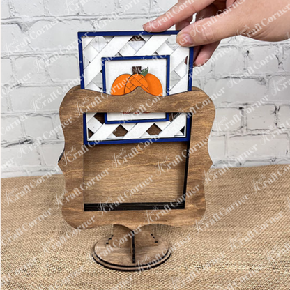 A hand places a pumpkin-themed card into the Heartland Drop In Swappable Squares Holder DIY Craft Kit (Set of 1), perfect for home décor. The scene shows a wooden surface and a white brick wall in the background.
