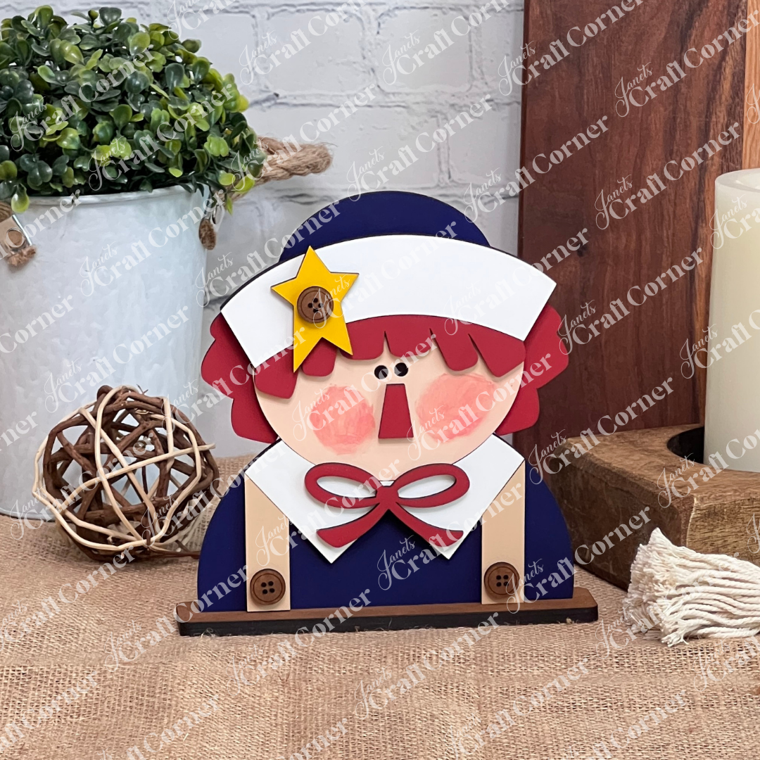 The Raggedy Ann & Andy Shelf Sitters DIY craft kit by Janet's Craft Corner features a red-haired wooden figure with a white bonnet, yellow star, buttons, and bow—sitting on burlap with rustic decor, perfect for fans and seasonal home decor.