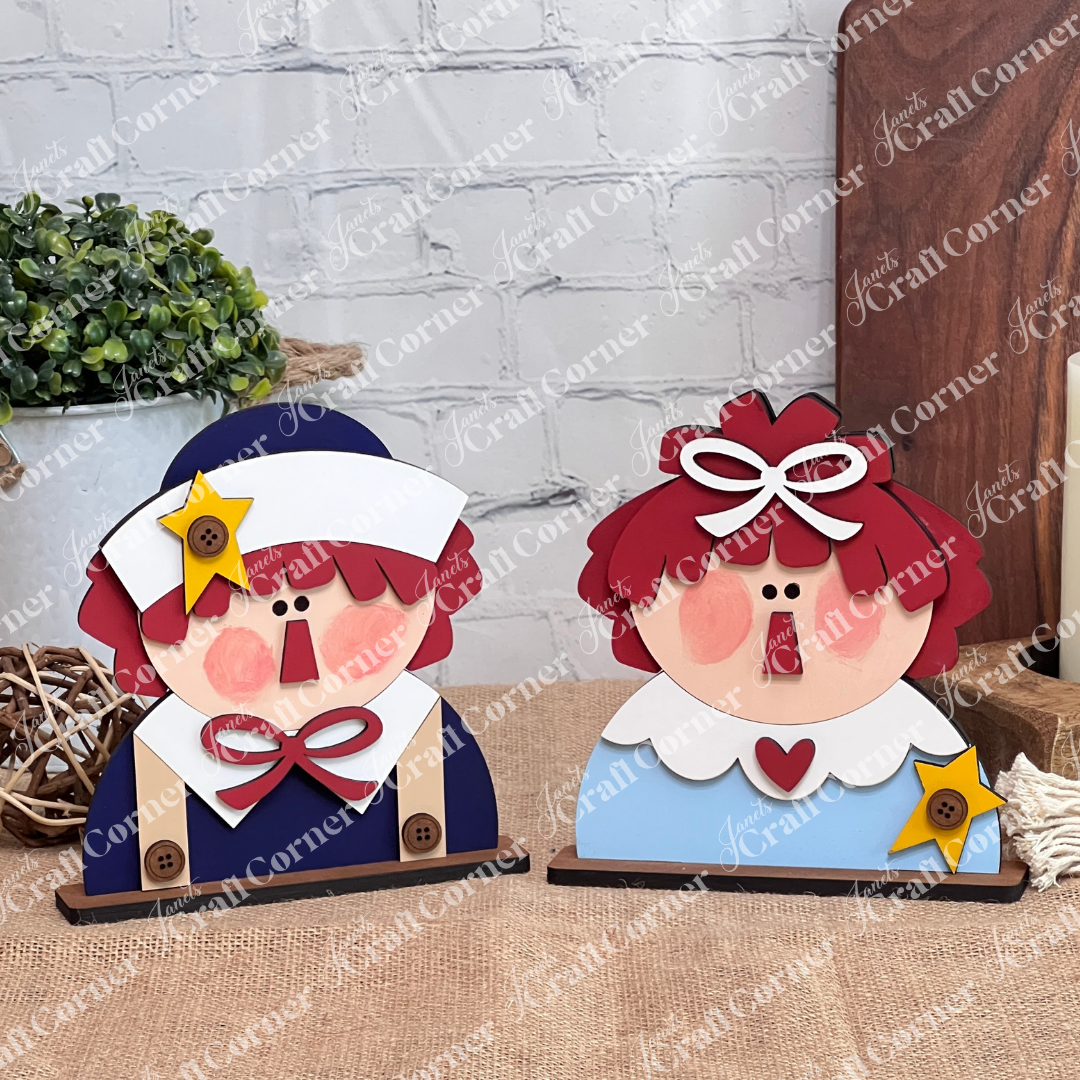 The Raggedy Ann & Andy Shelf Sitters DIY craft kit from Janet's Craft Corner features a set of 1 wooden figure with red yarn hair, rosy cheeks, and starry blue and beige outfits—ideal for adding seasonal charm to your home décor.