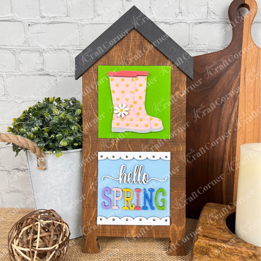 Janet's Craft Corner "Hello Spring Squares" is a wooden house-shaped sign with a pink rain boot and flower, plus "hello SPRING" on a colorful panel. Perfect for tiered tray decor with plants or candles!.