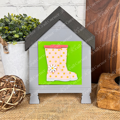 The Hello Spring Squares by Janet's Craft Corner is a gray house-shaped sign with a green swappable square, pink polka-dot rain boots, and a white daisy—ideal for tiered tray decor. Display it on a wooden surface with plants or woven accents.