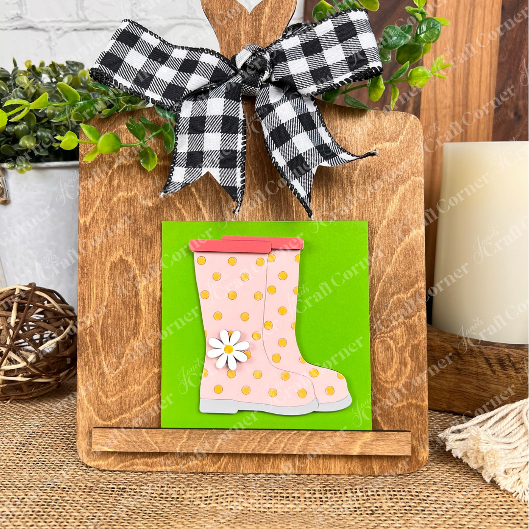Janet's Craft Corner Hello Spring Squares features a wooden display board with a checkered bow and a card showing a pink rain boot with gold dots and daisy on green—ideal for tiered tray decor. Greenery and candle add charm.