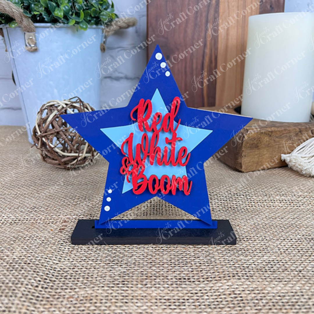 Janet's Craft Corner Patriotic Mini Shelf Sitters DIY Craft Kit includes a blue star with "Red White Boom" in red on a black base, perfect for Americana décor alongside candles, greenery, and rustic accents on burlap.