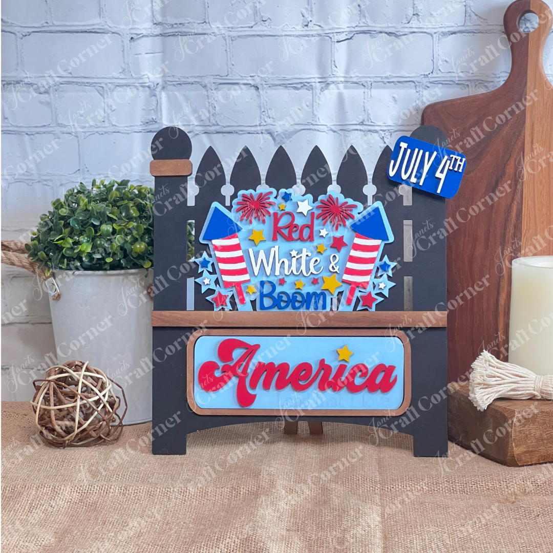 Janet's Craft Corner offers a "Red White & Boom Interchangeable Insert" featuring a black garden fence with fireworks and the phrases "Red White & Boom" and "America Land that I Love," plus a "July 4th" license plate, all on a white brick background—perfect for DIY seasonal home decor.