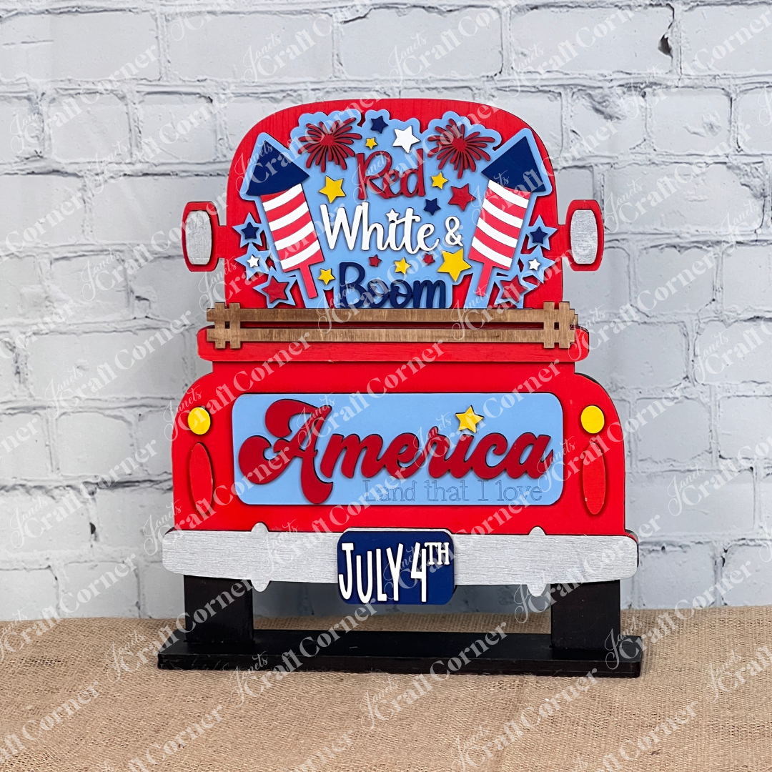 Janet's Craft Corner offers a "Red White & Boom Interchangeable Insert" featuring a patriotic red truck with fireworks and the phrases "Red White & Boom" and "America Land that I Love," plus a "July 4th" license plate, all on a white brick background—perfect for DIY seasonal home decor.