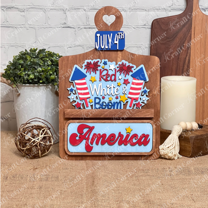 Janet's Craft Corner offers a "Red White & Boom Interchangeable Insert" featuring a stained breadboard with fireworks and the phrases "Red White & Boom" and "America Land that I Love," plus a "July 4th" license plate, all on a white brick background—perfect for DIY seasonal home decor.
