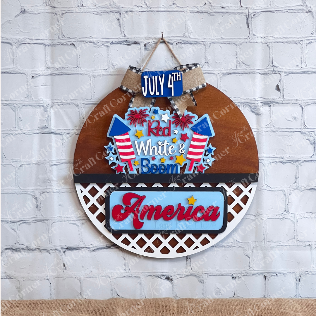 The Janet's Craft Corner "Red White & Boom" Interchangeable Insert is a DIY home decor piece featuring "July 4th" on a small sign and "Red, White, & Boom" with fireworks on the main sign. Below, it states "America and that I love," set against a lattice background with a white brick wall backdrop.
