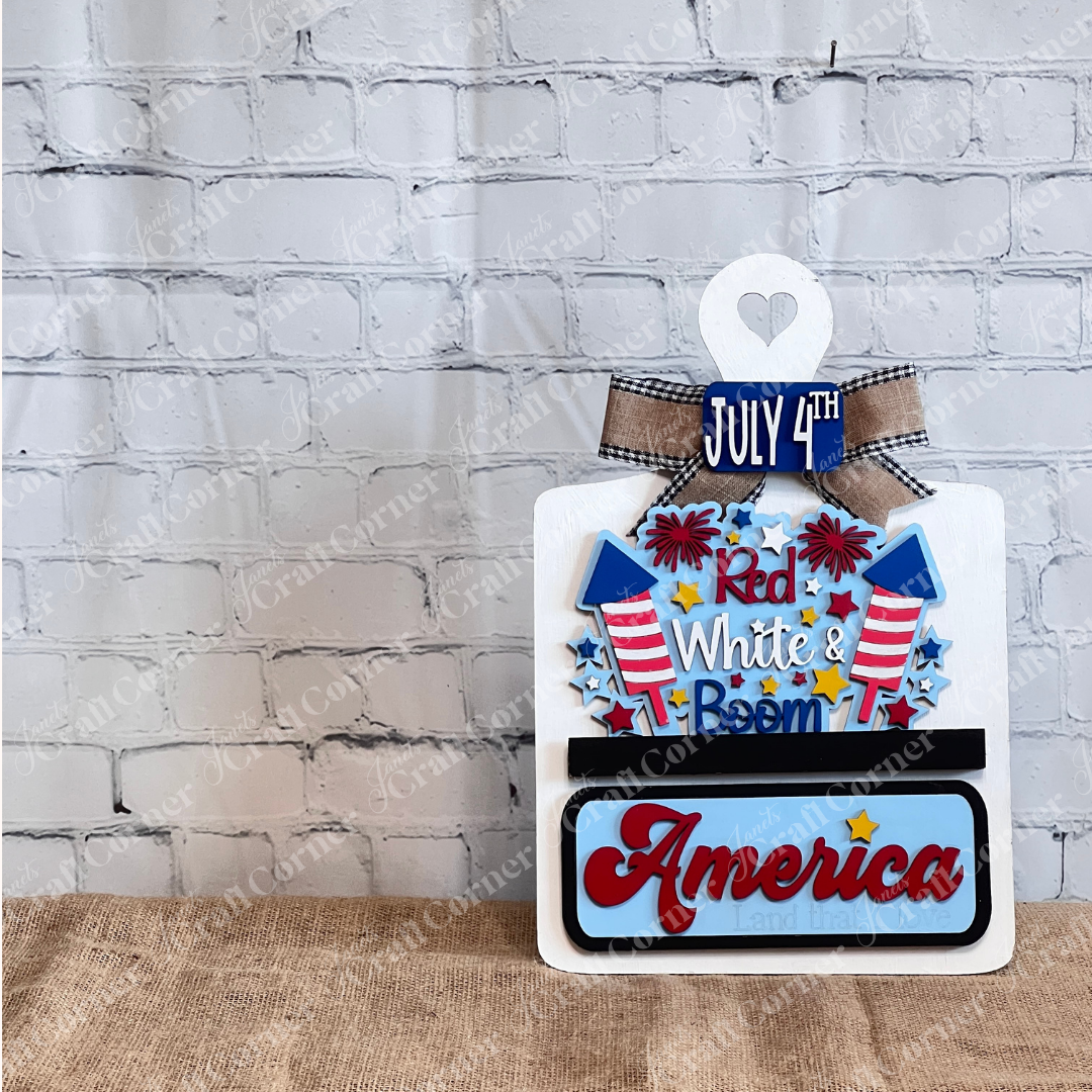 The Red White & Boom Interchangeable Insert from Janet's Craft Corner is a DIY home decor craft kit that features "July 4th," fireworks, and "Red White & Boom" in patriotic colors along with “America, Land that I Love” against a white brick wall backdrop.