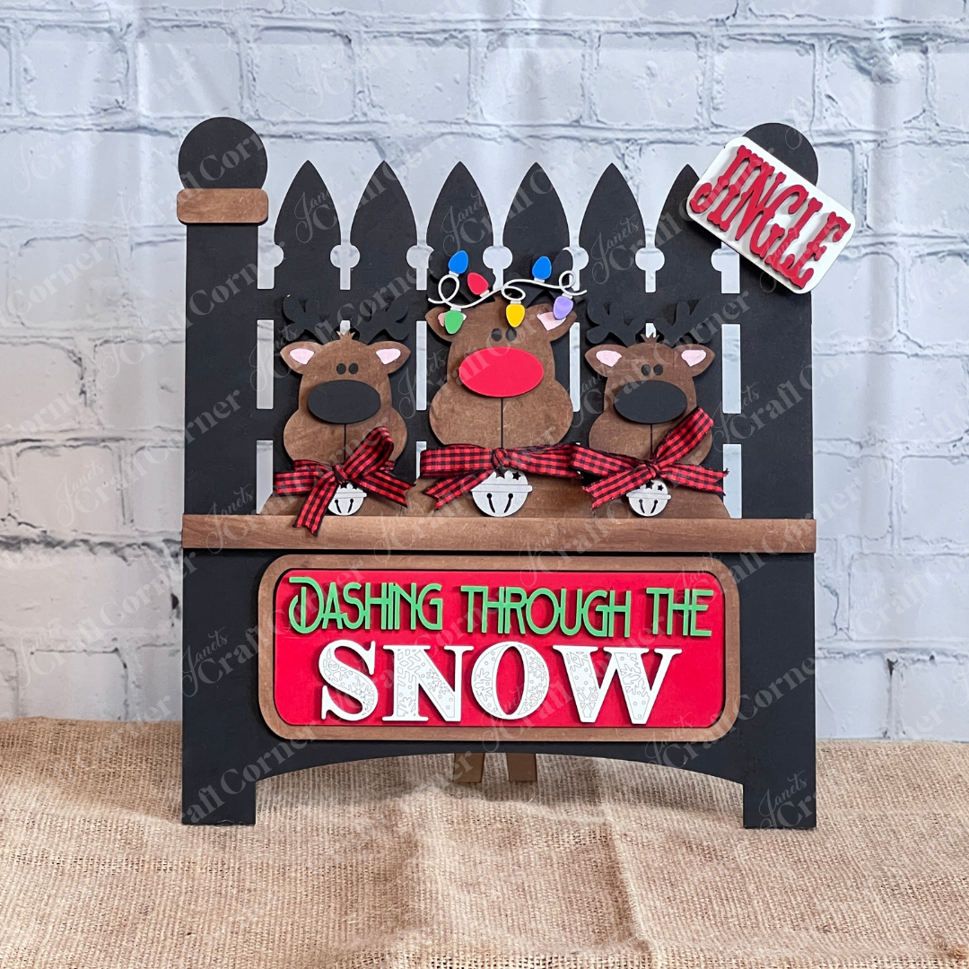 Janet's Craft Corner offers a Garden Fence with Insert DIY kit featuring three cartoon reindeer with red noses and plaid scarves. It includes "Jingle" and "Dashing Through the Snow" signs, interchangeable inserts, and colorful lights for festive charm.