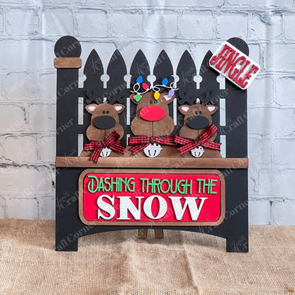Janet's Craft Corner offers the Reindeer Interchangeable Insert, a DIY seasonal home decor kit featuring a black garden fence with cartoon reindeer and festive text against a white brick wall on burlap—ideal for hand-painted décor enthusiasts.