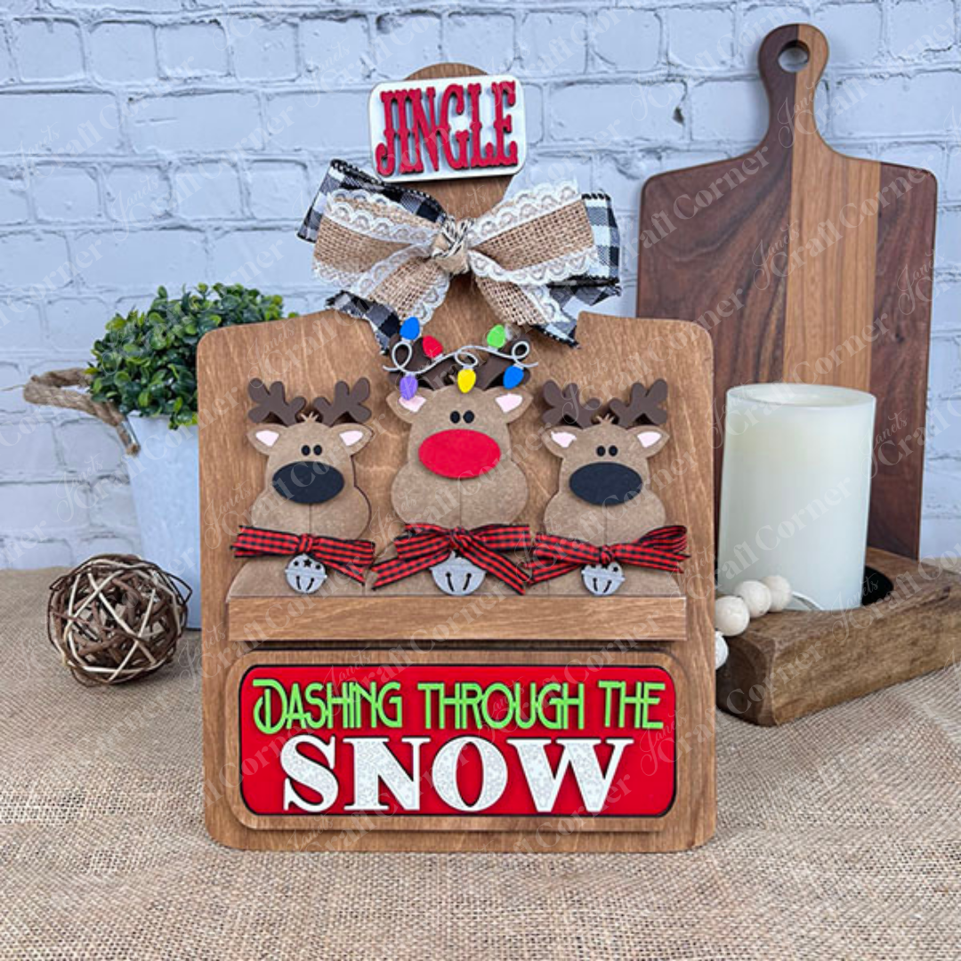 Janet's Craft Corner presents the Breadboard Interchangeable Base with Insert. This DIY home decor kit includes a wooden holiday piece featuring three reindeer with plaid bows, a "Jingle" sign, and "Dashing Through the Snow," complemented by accessories like a candle, plant, and decorative ball.