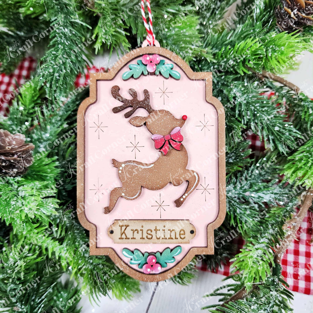 The North Pole Ornaments DIY craft kit by Janet's Craft Corner features a reindeer with a red bow, pink background, floral accents, and "Kristine"—a perfect festive decor or holiday crafting inspiration. Set of 1.