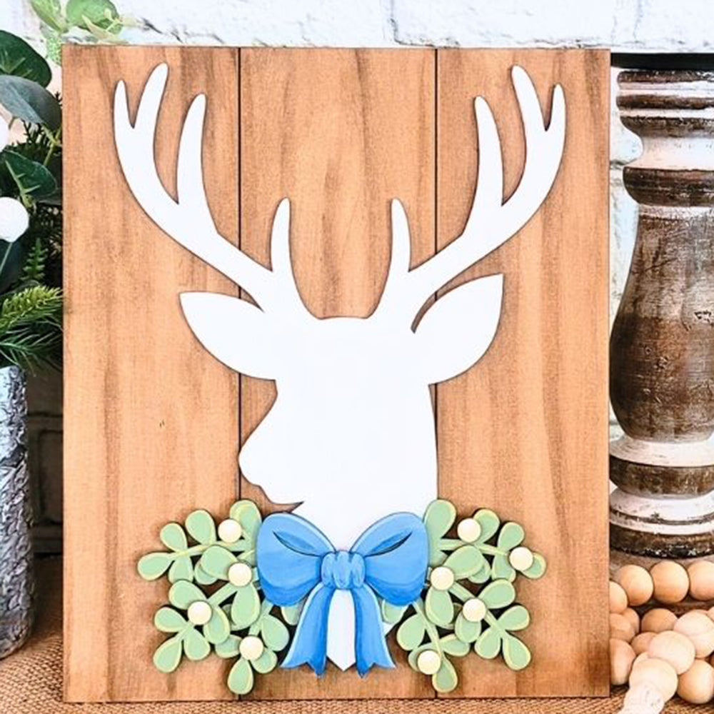 The Rustic Deer Shelf Leaner DIY Home Decor Kit by Janet's Craft Corner offers a delightful piece of festive decor. It includes a wooden plaque adorned with a white silhouette of a deer’s head and antlers, accented by green leaves and yellow berries below a blue bow. The setup is completed with a rustic vase surrounded by natural greenery.