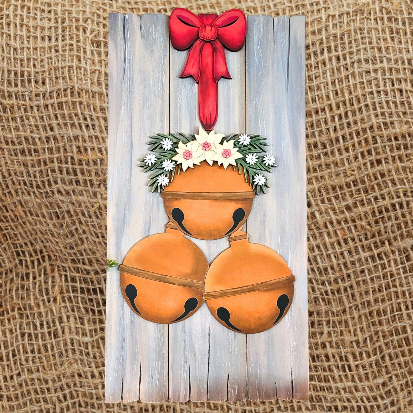 Introducing the Rustic Bell Cluster Leaner DIY Home Decor Kit from Janet's Craft Corner, featuring three brown jingle bells embellished with white flowers and greenery. These charming elements dangle against a reclaimed barn wood backdrop, while a red bow adds a touch of elegance to its rustic beauty, all set against textured burlap.