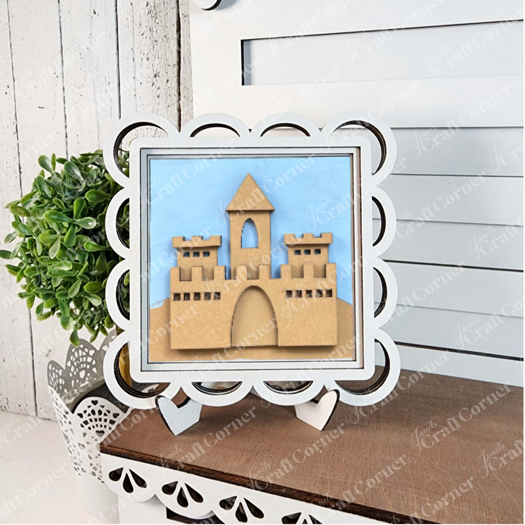 The "Summer Beach Squares" DIY craft kit from Janet's Craft Corner features framed sandcastle art with a blue background, styled on a white stand beside greenery—ideal seasonal decor for small spaces or tiered tray displays.