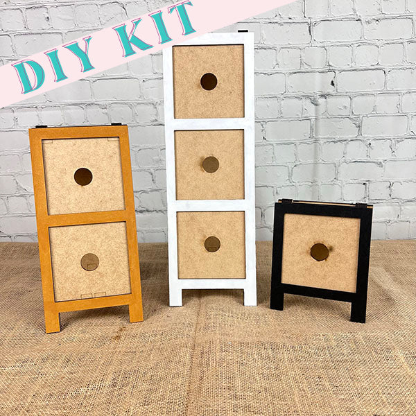 Introducing the Sandwich Boards Interchangeable Square Holders from Janet's Craft Corner, a craft kit featuring three miniature wooden storage units with circular handles. Set against a white brick wall, these units are available in three colors: natural wood with three drawers, white with three drawers, and black with one drawer. Enjoy versatile designs that can adapt to any decor style.